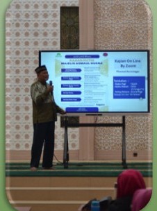 Foto dokumentasi: seorang pengajar berdiri dan berbicara di depan layar presentasi kajian daring di ruang masjid.